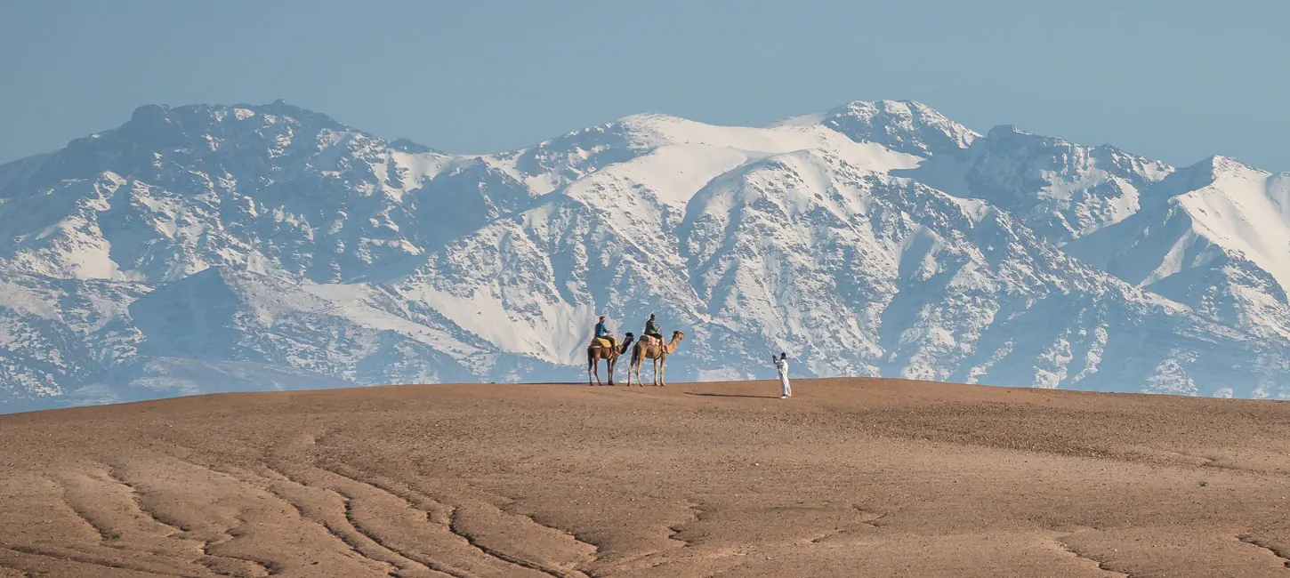 Deux voyageurs à dos de dromadaire face aux montagnes enneigées de l’Atlas